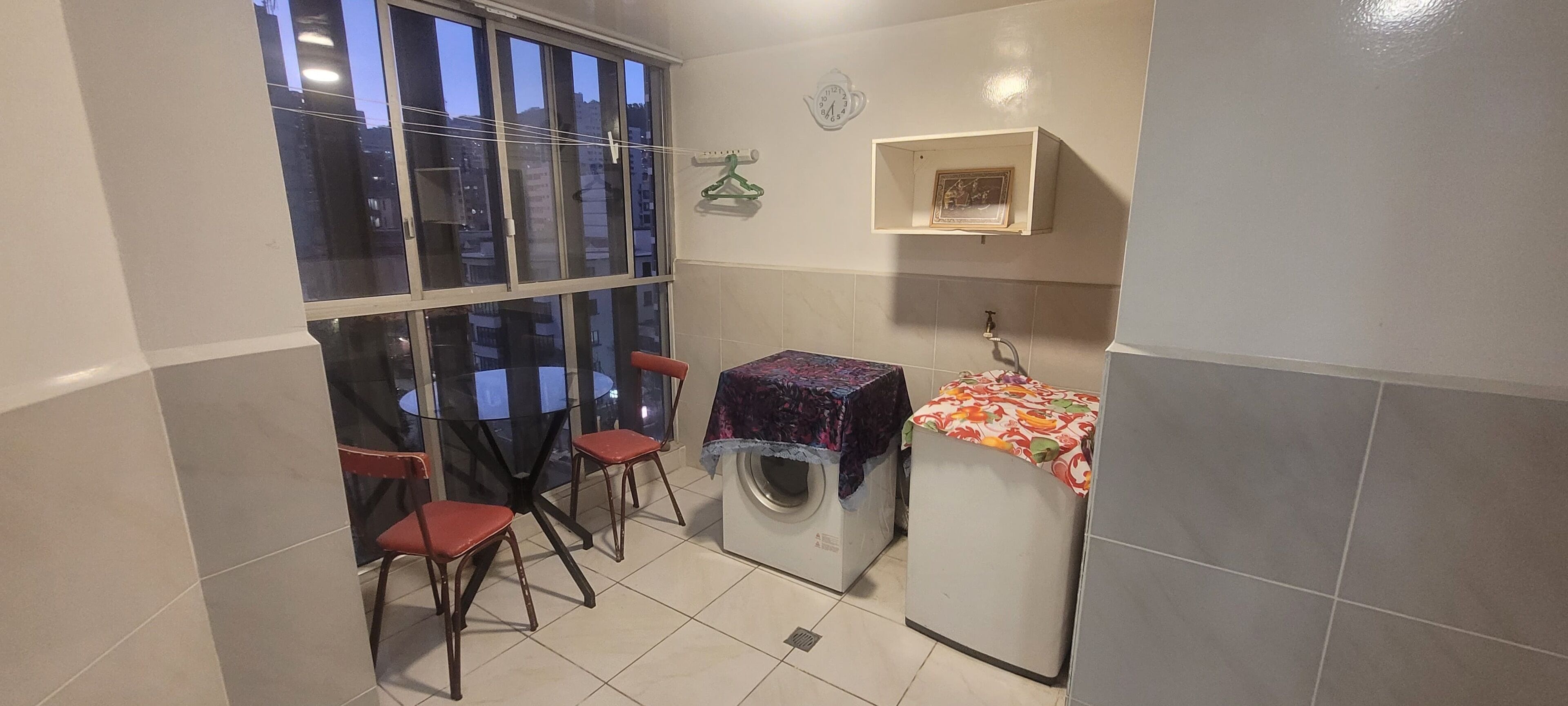Une petite salle de bain équipée d’un lave-linge, d’une petite table recouverte d’une nappe, de deux chaises et d’une fenêtre donnant sur des immeubles.