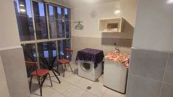 Une petite salle de bain équipée d’un lave-linge, d’une petite table recouverte d’une nappe, de deux chaises et d’une fenêtre donnant sur des immeubles.