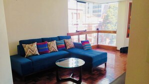 Una sala de estar con un sofá azul, almohadones coloridos, una mesa de centro de vidrio y suelo de madera.