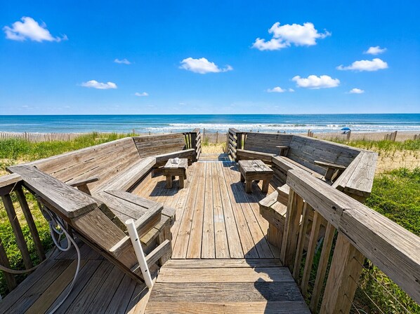 A wooden deck with benches and tables, leading to a beach with the ocean in the background.