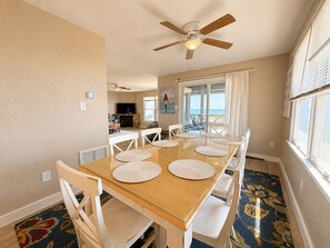 A dining area with a wooden table and white chairs, a ceiling fan, and a view of the ocean through a sliding glass door. - A Sense of Porpoise - 4 BR Oceafront Townhome in Southern Shores (Southern Shores)