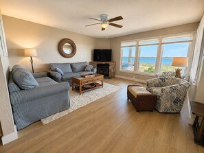 Una sala de estar con sofá, sillón, mesa de centro, chimenea, televisor y ventanas amplias con vistas.