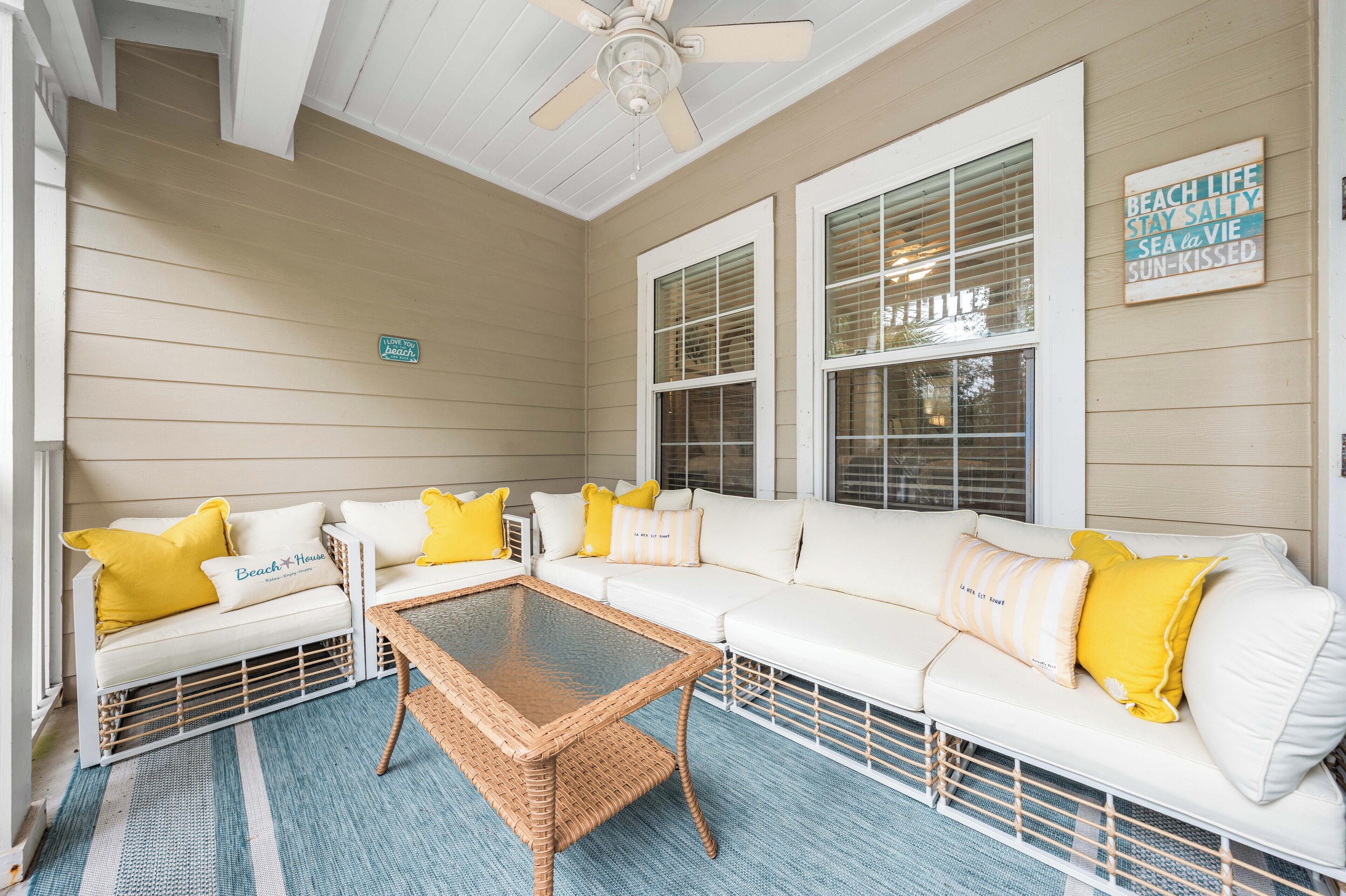 A covered porch with white sofas, yellow pillows, and a wicker table.