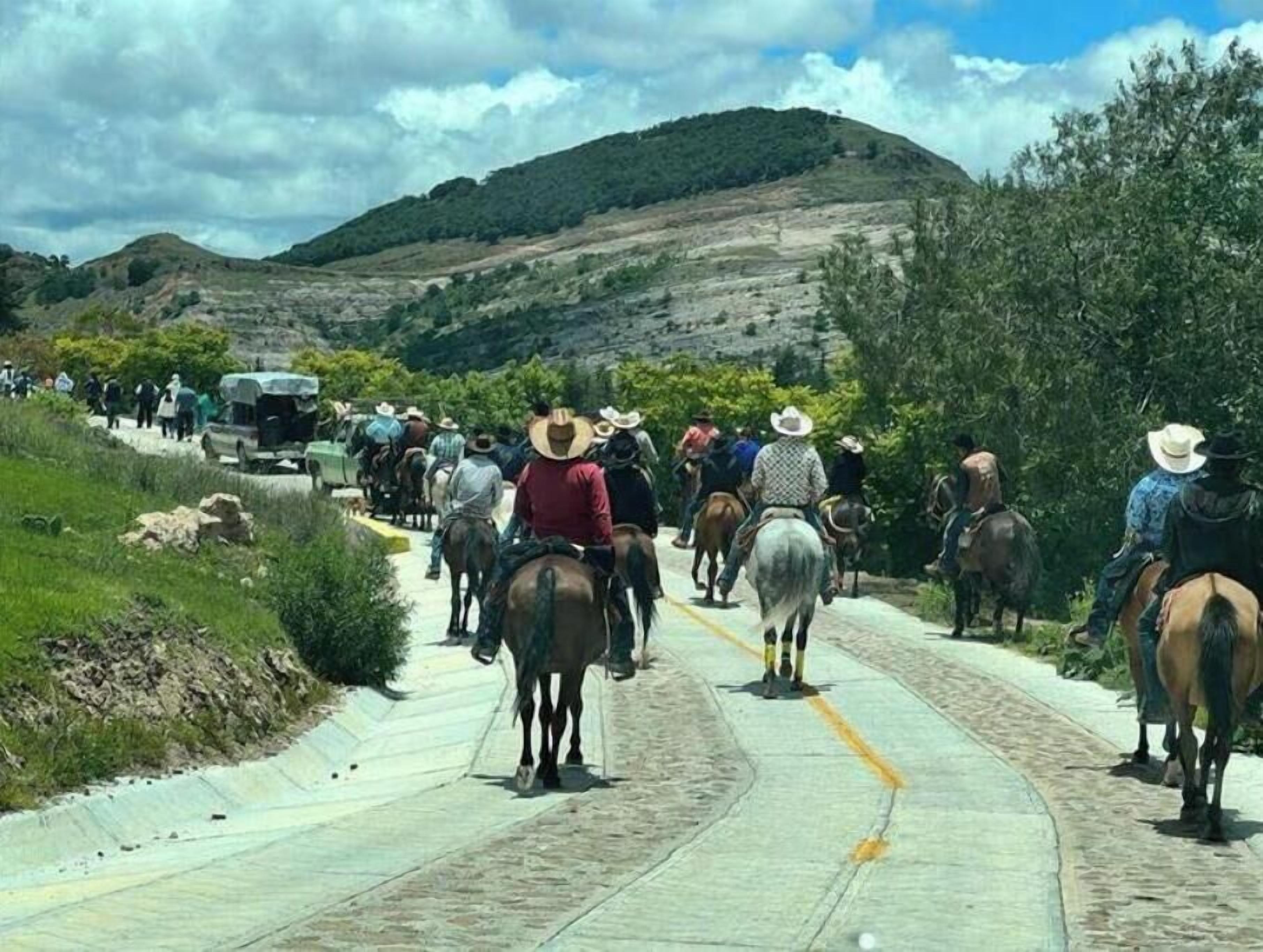Eine Gruppe von Personen auf Pferden auf einer asphaltierten Straße mit einer Berglandschaft im Hintergrund.