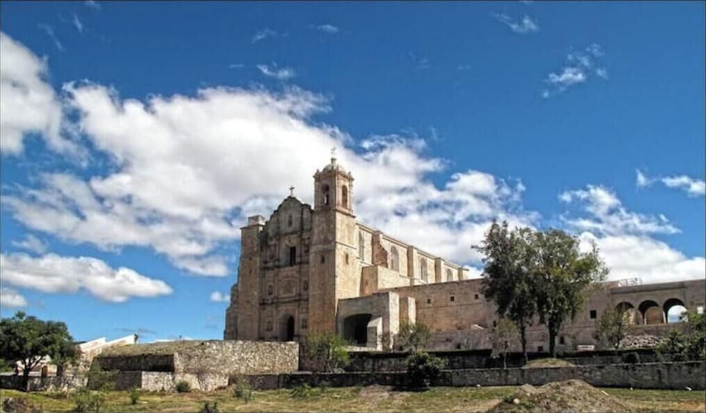 En historisk byggnad med ett framträdande klocktorn och välvda fönster, omgiven av en stenmur och grönska.