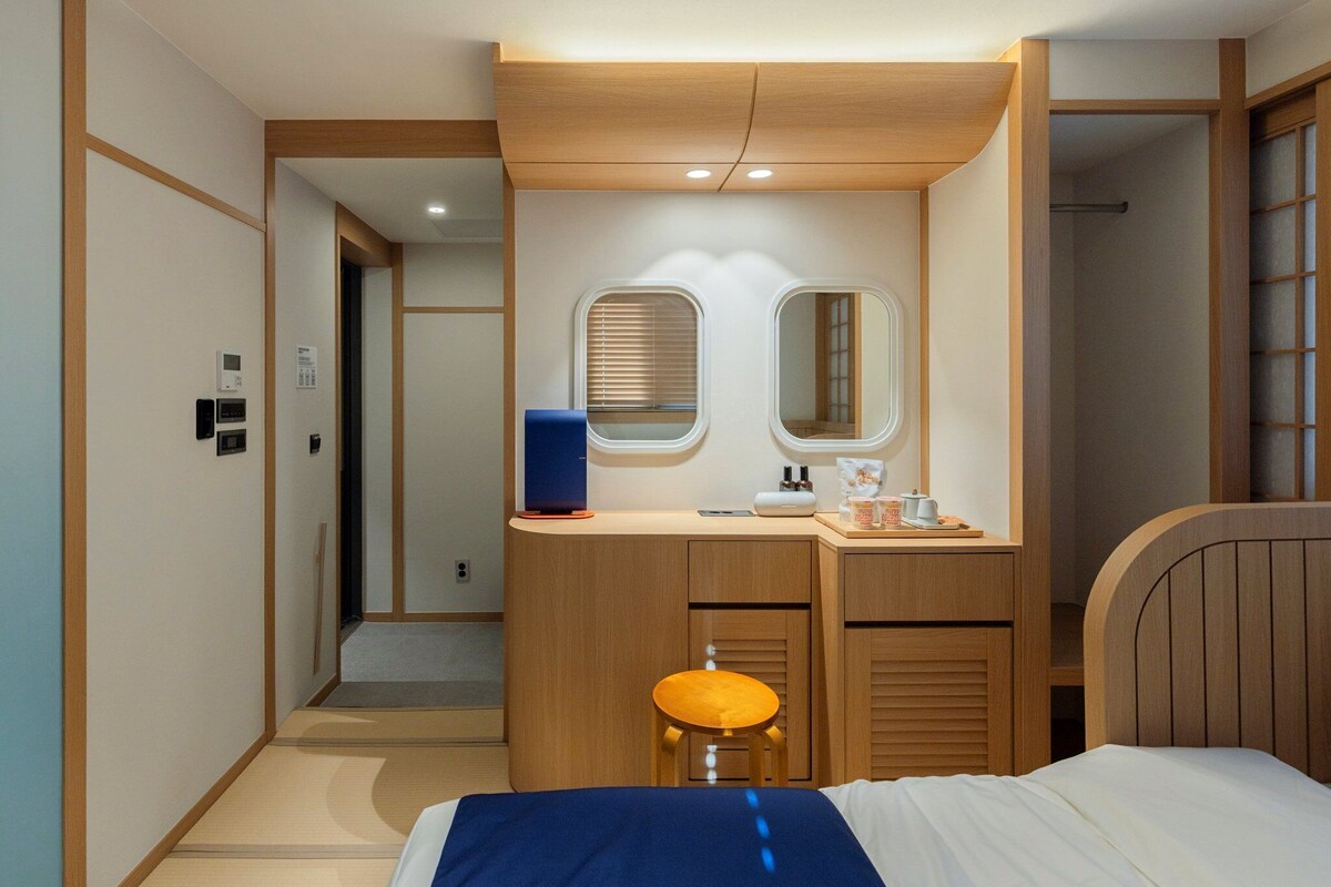 A modern hotel room with a wooden bed, a desk with a chair, a mirror, and a small stool.