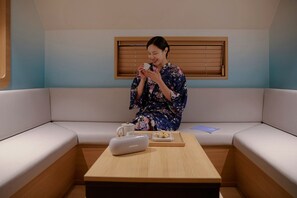 En person sidder på en sofa i et rum med et træbord, et vindue med persienner og en lille bakke med kop og snacks.