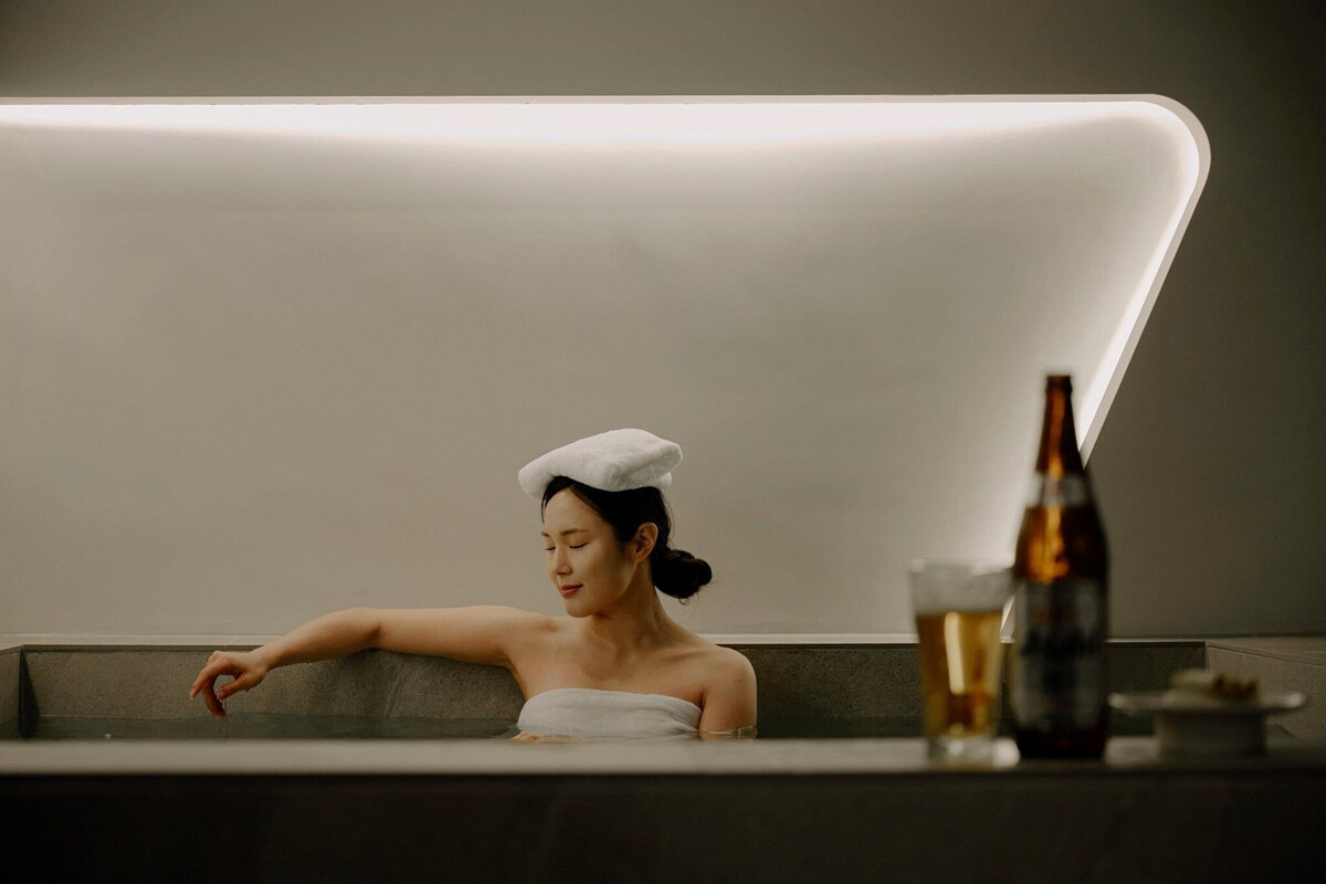 A person in a bathtub with a towel on their head, a bottle, and a glass on a table.