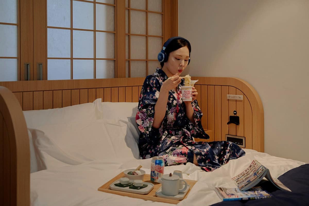 A person sitting on a bed with a tray of food, headphones, and a magazine.