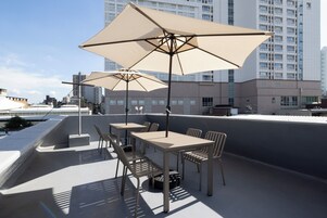 Un espacio al aire libre con mesas y sillas, sombrillas grandes y vistas a un paisaje urbano con edificios altos.