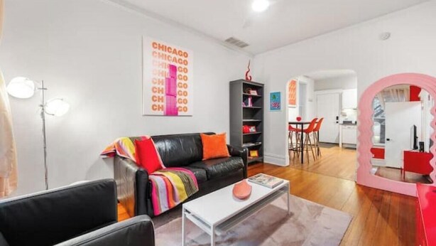 A modern living room with a black leather sofa, a white coffee table, and a colorful throw blanket. There is a bookshelf and a dining area with a pink mirror. - Chicago Citrus Charm 2BR  WestTown (Chicago)