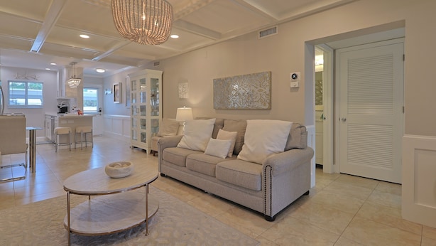 A modern living room with a beige sofa, a coffee table, and a chandelier. There is a kitchen area in the background with a dining table and chairs.