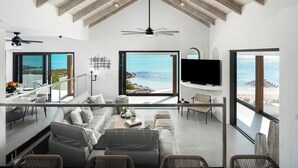 A spacious living area with a high ceiling, large windows offering ocean views, a flat-screen TV, and a dining area with a glass railing. - Opaline Villa -3BR 2bath Oceanfront (Providenciales)