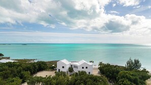 Un bâtiment blanc doté d’un toit incurvé, entouré d’une végétation luxuriante, qui surplombe une mer turquoise sous un ciel dégagé.