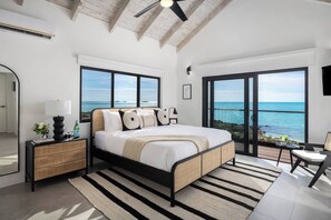 Une chambre avec un grand lit, une table de chevet, une chaise, une vue sur l’océan et un ventilateur de plafond.