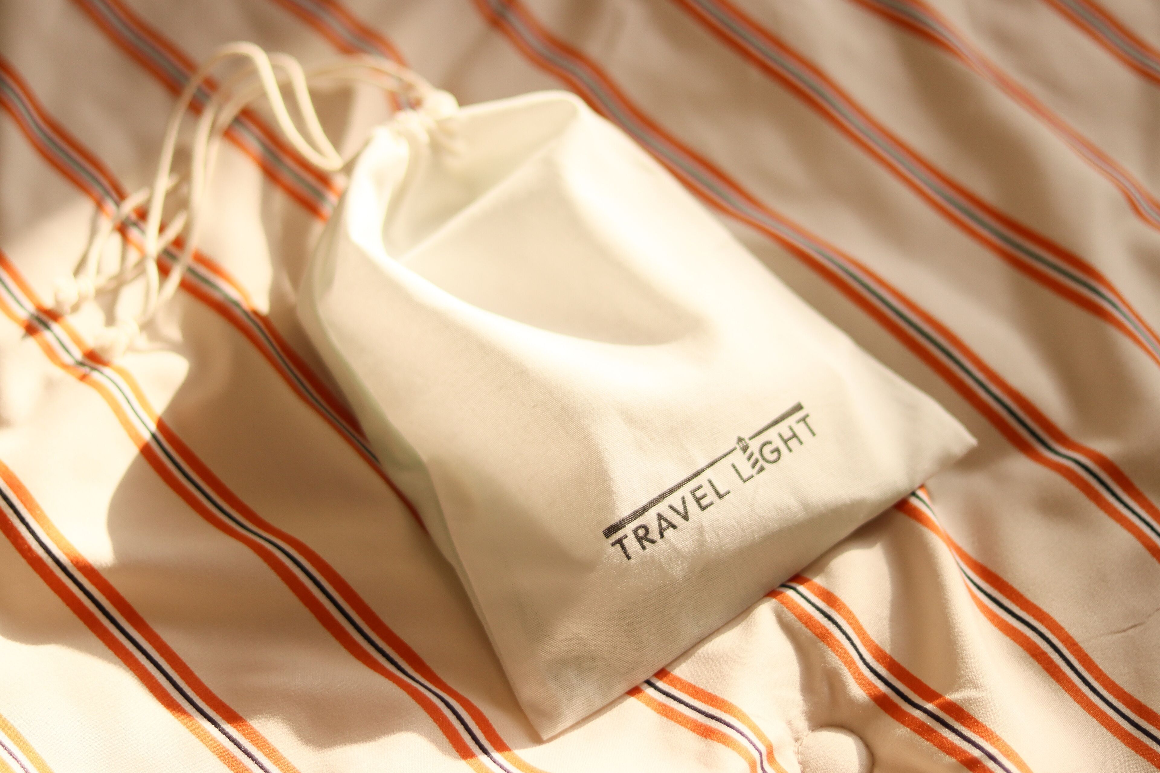A white drawstring bag with "TRAVEL LIGHT" printed on it, placed on a striped fabric.