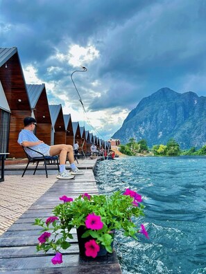 A wooden deck with a person fishing, overlooking a body of water and mountains. - Heun Phea Meuang Feuang Riverside2 (Muang Fuang)