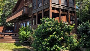 Une cabane en bois à deux étages, dotée d’un porche couvert et d’une petite terrasse, entourée d’une végétation luxuriante.