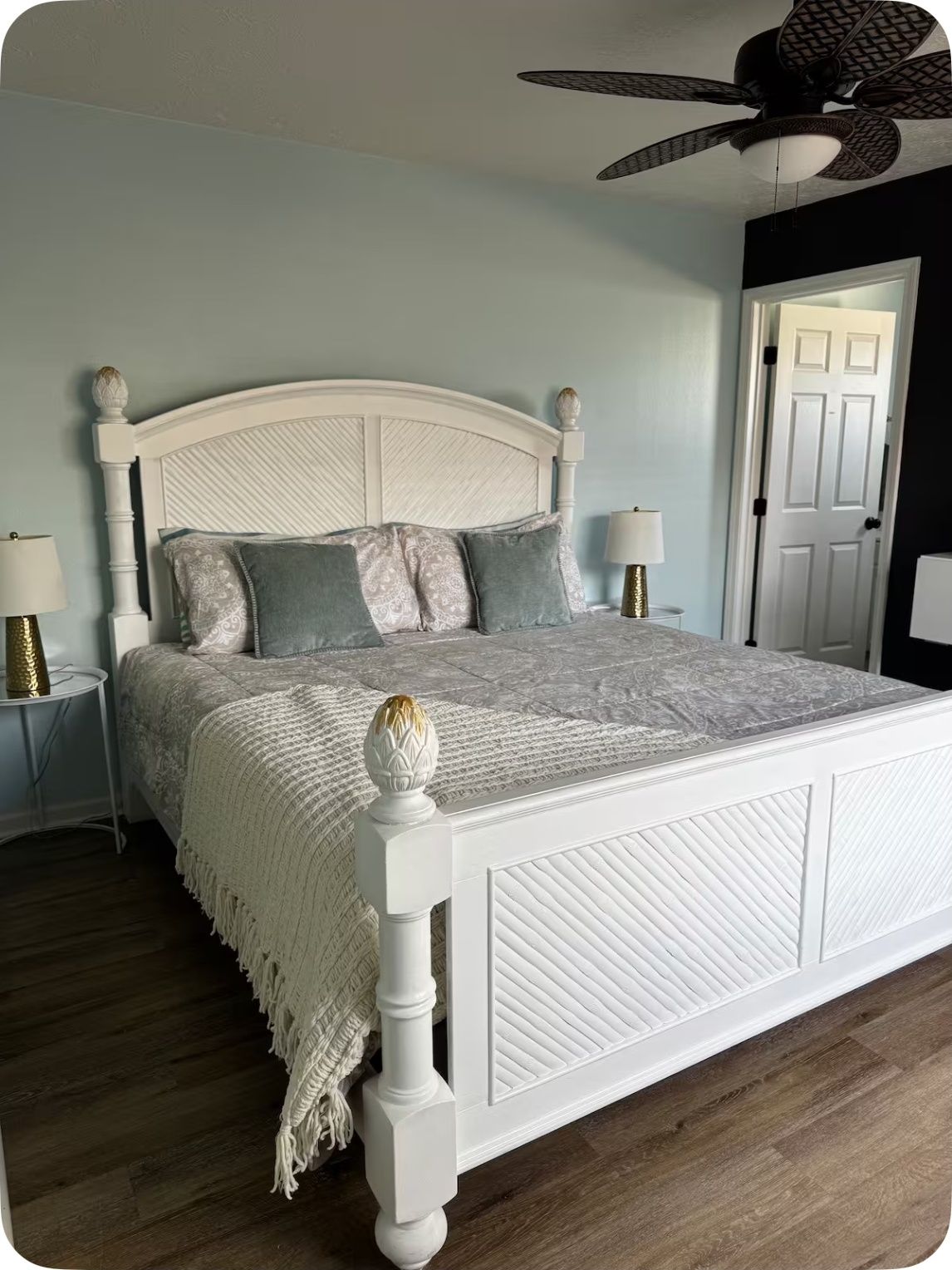Un dormitorio con cama blanca, almohadas estampadas, ventilador de techo y una puerta al fondo.
