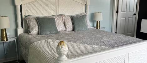 Un dormitorio con cama blanca, almohadas estampadas, ventilador de techo y una puerta al fondo.