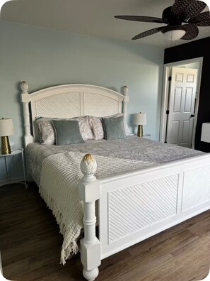 Un dormitorio con cama blanca, almohadas estampadas, ventilador de techo y una puerta al fondo.