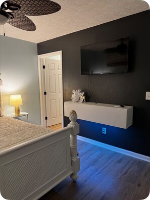 Une chambre à coucher comprenant un lit, une lampe de chevet, un téléviseur fixé au mur et une porte donnant sur une autre pièce.
