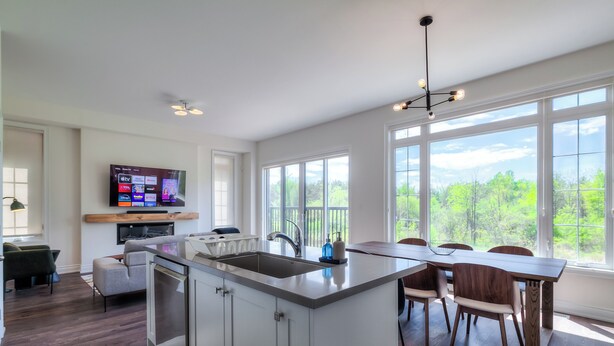 Una cocina moderna con una isla central, un comedor con mesa y sillas de madera, y un amplio ventanal con vistas al verdor.