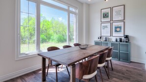 Un comedor con mesa y sillas de madera, ventanas amplias con vista a la vegetación y una zona de cocina con armarios y obras de arte enmarcadas.