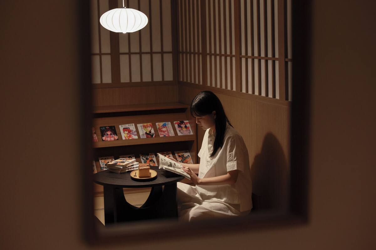 A person sitting at a table reading a book in a room with wooden walls and a hanging light fixture.