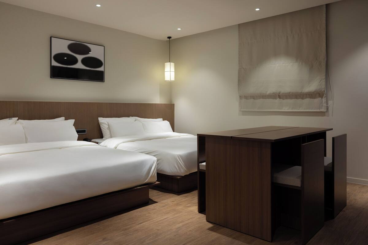 A hotel room with two beds, a wooden desk, and a wall-mounted light fixture.