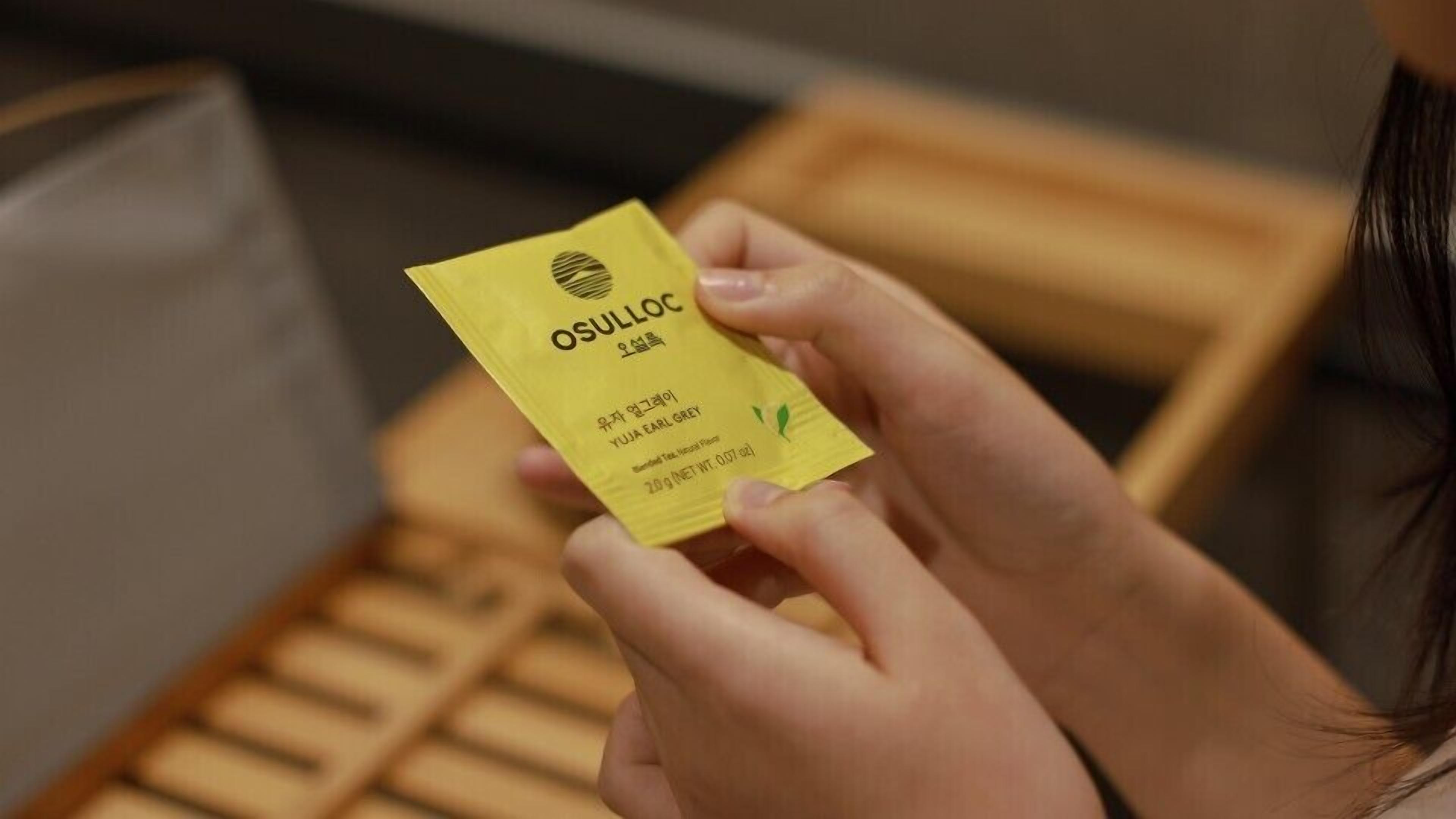 Une personne tient un paquet jaune portant l’inscription « OSULLOC », avec du texte dans une autre langue, et l’inscription « YUJA EARL GREY ».