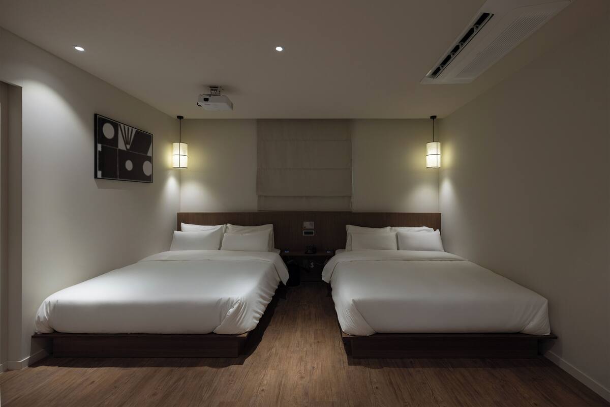 A hotel room with two beds, wooden flooring, recessed ceiling lights, a projector, and wall-mounted lamps.