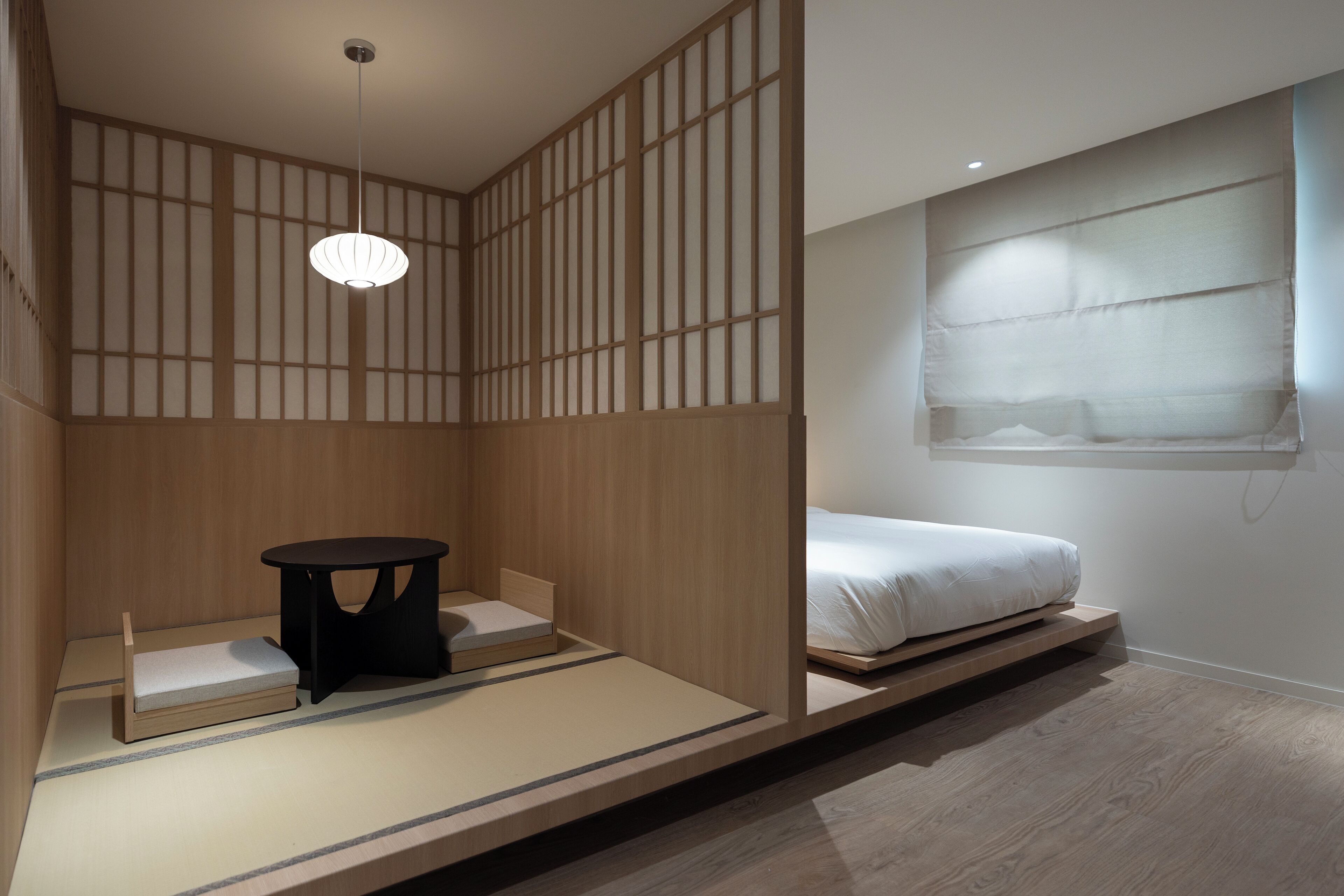 A minimalist Japanese-style room with tatami flooring, a low platform bed, a small black table, and a hanging light fixture.