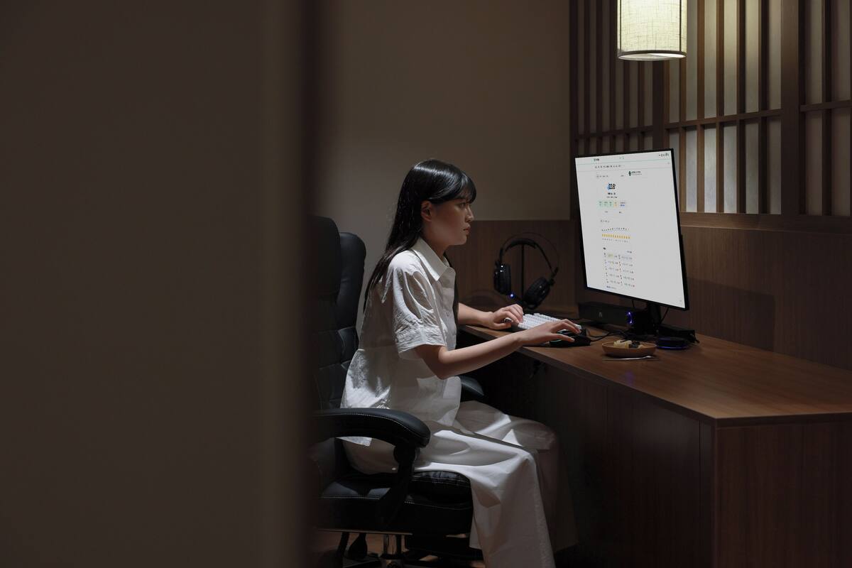A person seated at a desk with a computer, headphones, and a lamp.