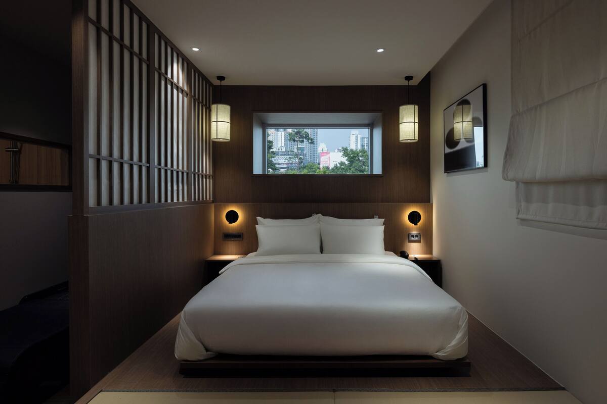 A modern hotel room with a large bed, two bedside tables with lamps, a window with a city view, and a wall-mounted artwork.