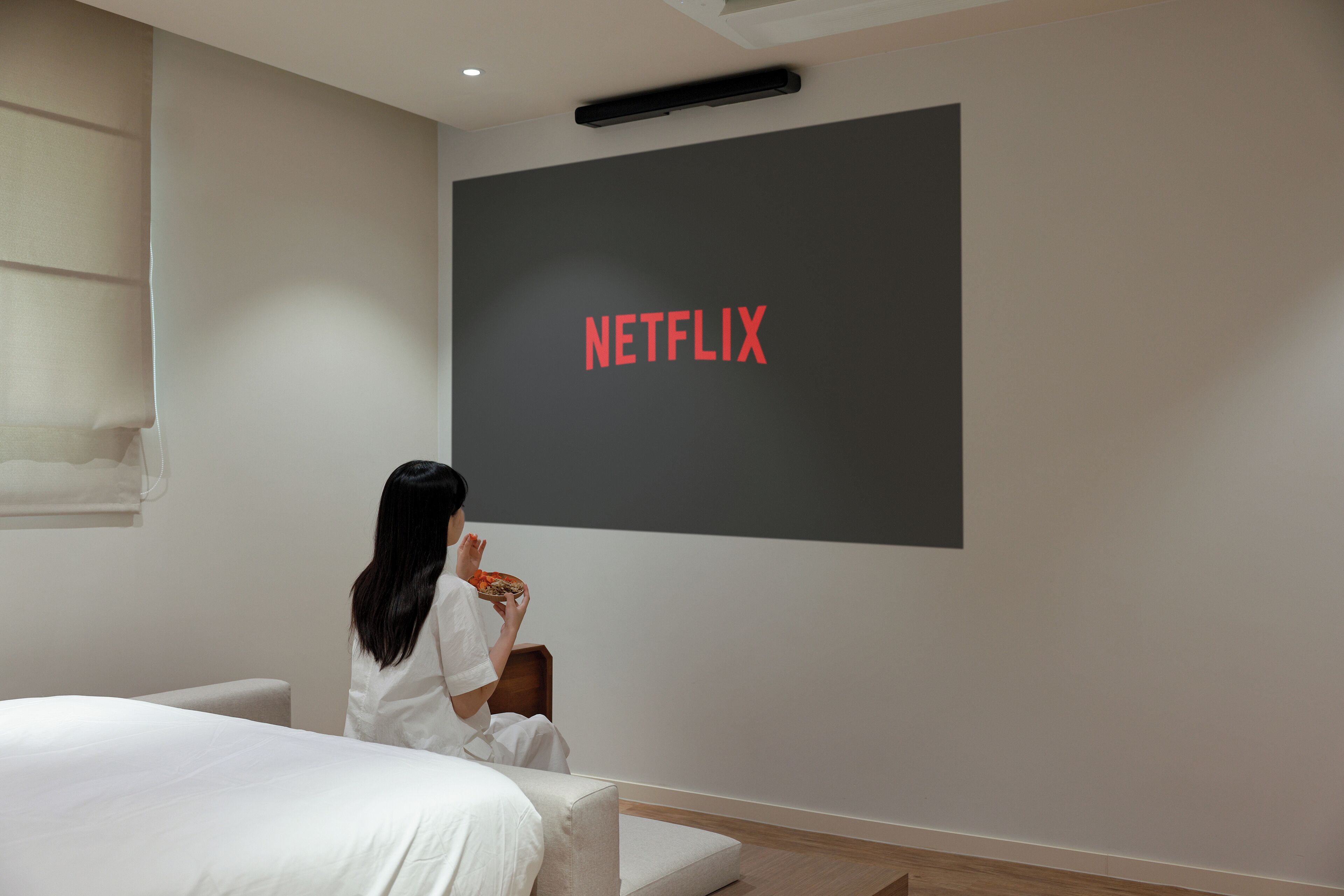 Ein Zimmer mit einem Bett, einem an der Wand befestigten Bildschirm, der das Wort 'NETFLIX' zeigt, und einer Person, die auf einem Stuhl sitzt.