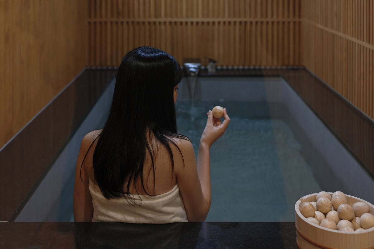 A person sitting in a wooden bath with a bowl of wooden balls beside them.
