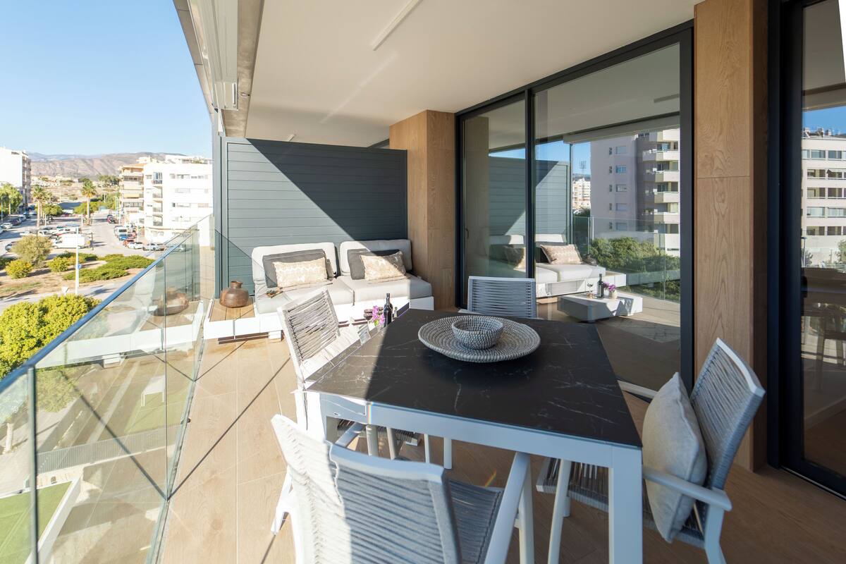 Un balcón con barandilla de vidrio, una mesa con sillas y vista a la ciudad.