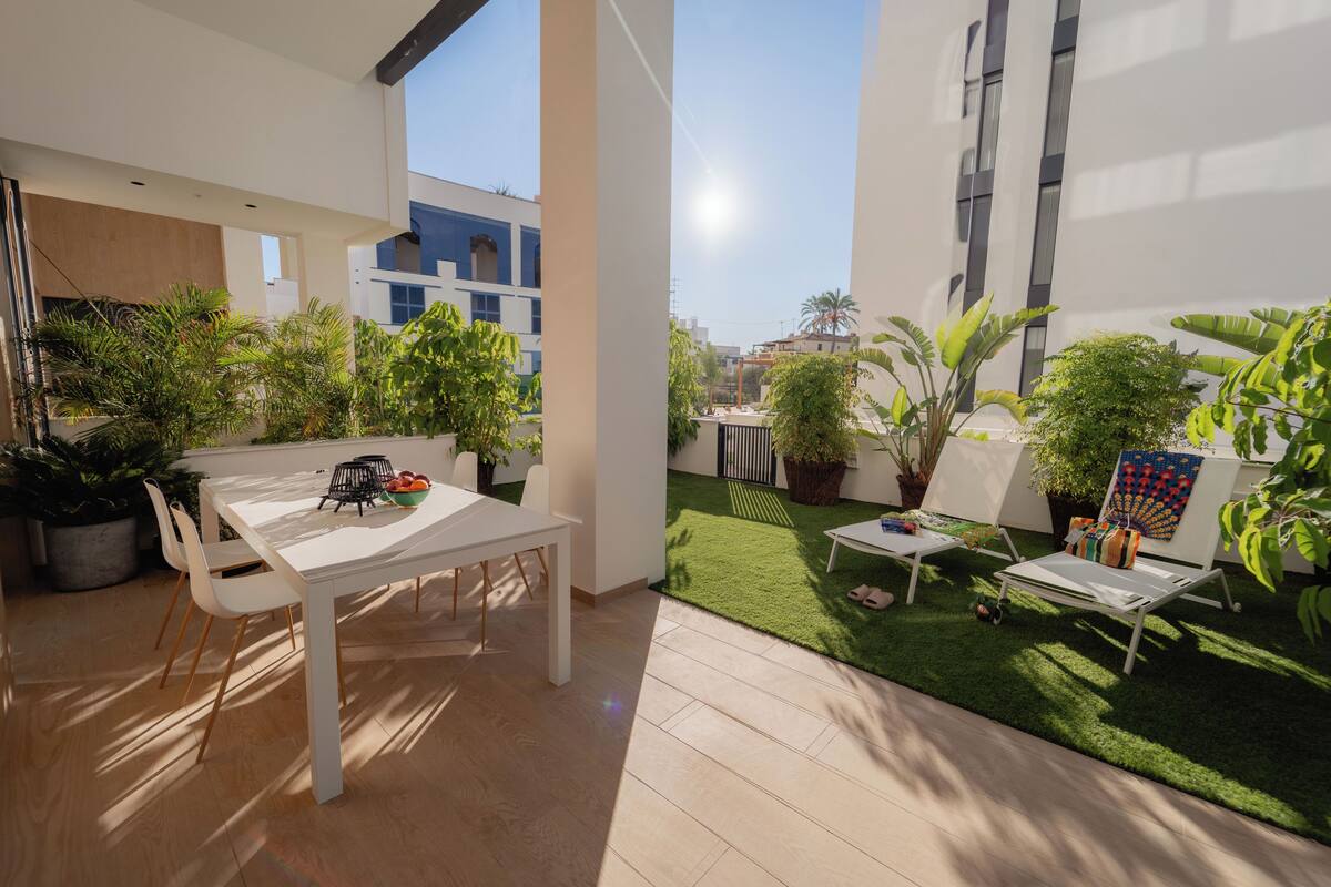 Un patio moderno al aire libre con una mesa de comedor, sillas y un pequeño jardín con césped artificial y plantas en macetas.