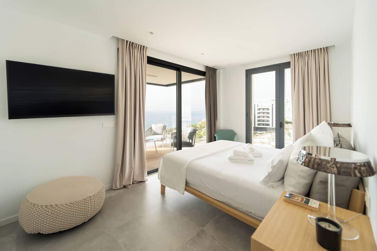 Una habitación de hotel moderna con una cama grande, un televisor de pantalla plana montado en la pared y vistas a la ciudad a través de ventanas amplias.