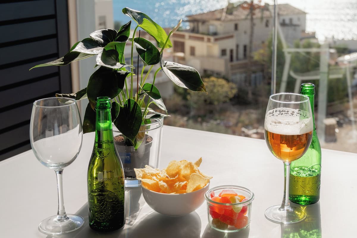 Una mesa con una botella verde, una copa de vino, un bol de patatas fritas y una cerveza, con vistas al mar y edificios al fondo.