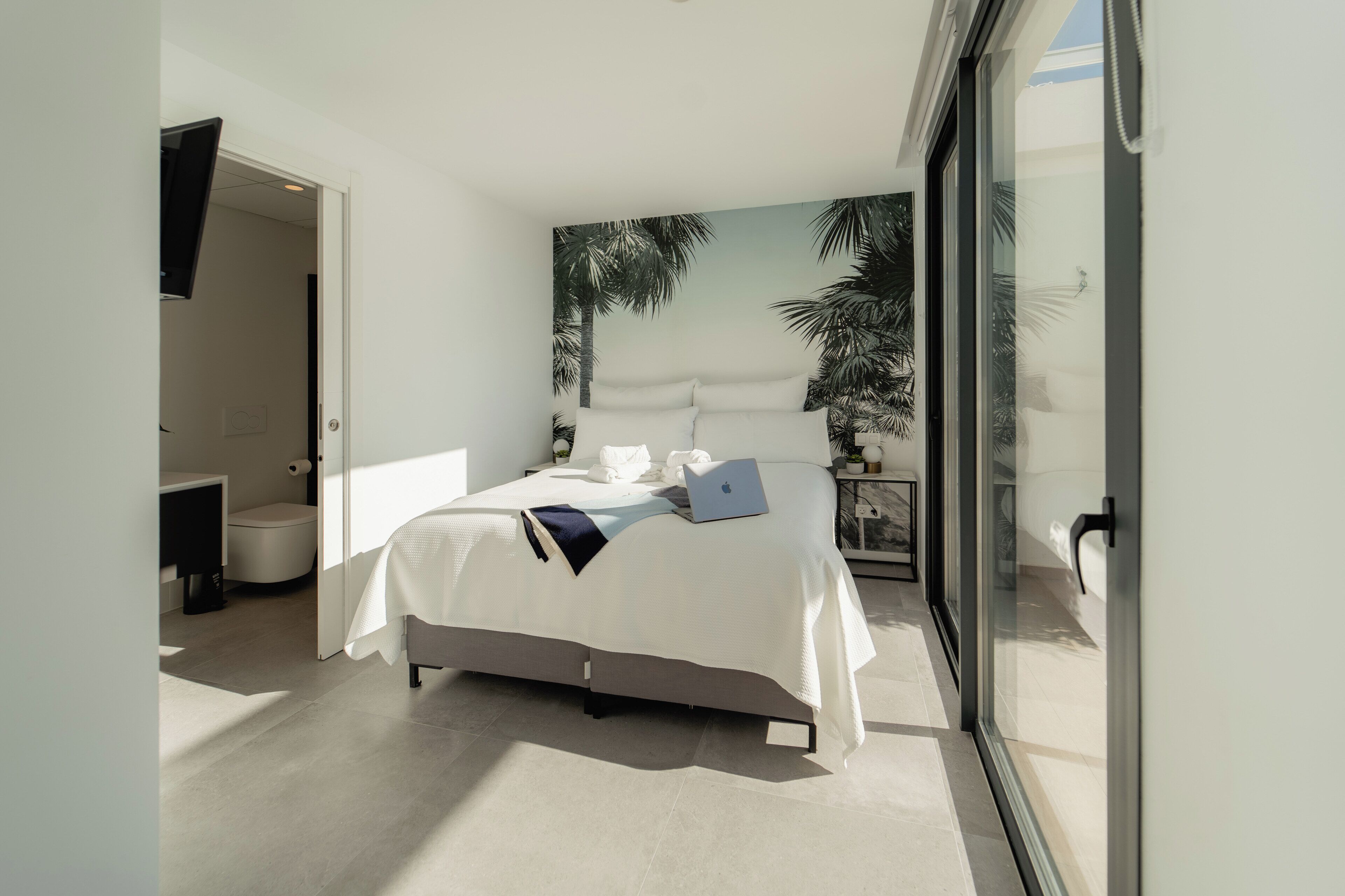 Un dormitorio moderno con una cama grande, una computadora portátil y vistas a palmeras a través de la ventana.