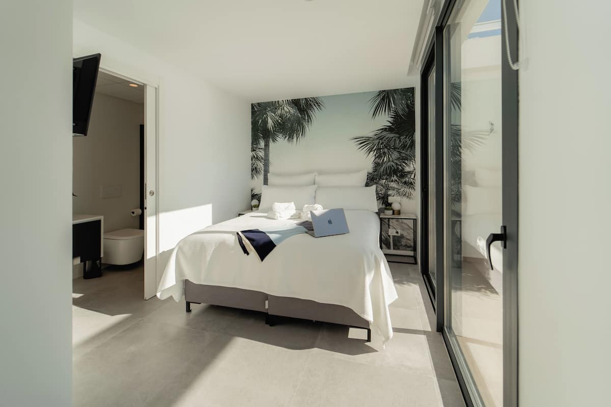 Un dormitorio moderno con una cama, una computadora portátil y un ventanal con vistas a palmeras.