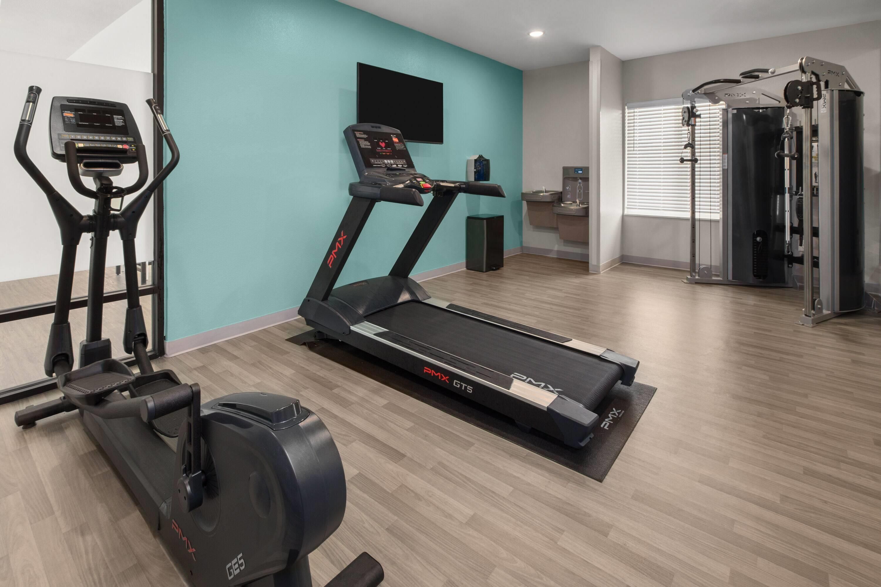 Ein Fitnessraum mit Cardiogeräten, einem an der Wand montierten Fernseher und einer Trinkstation.