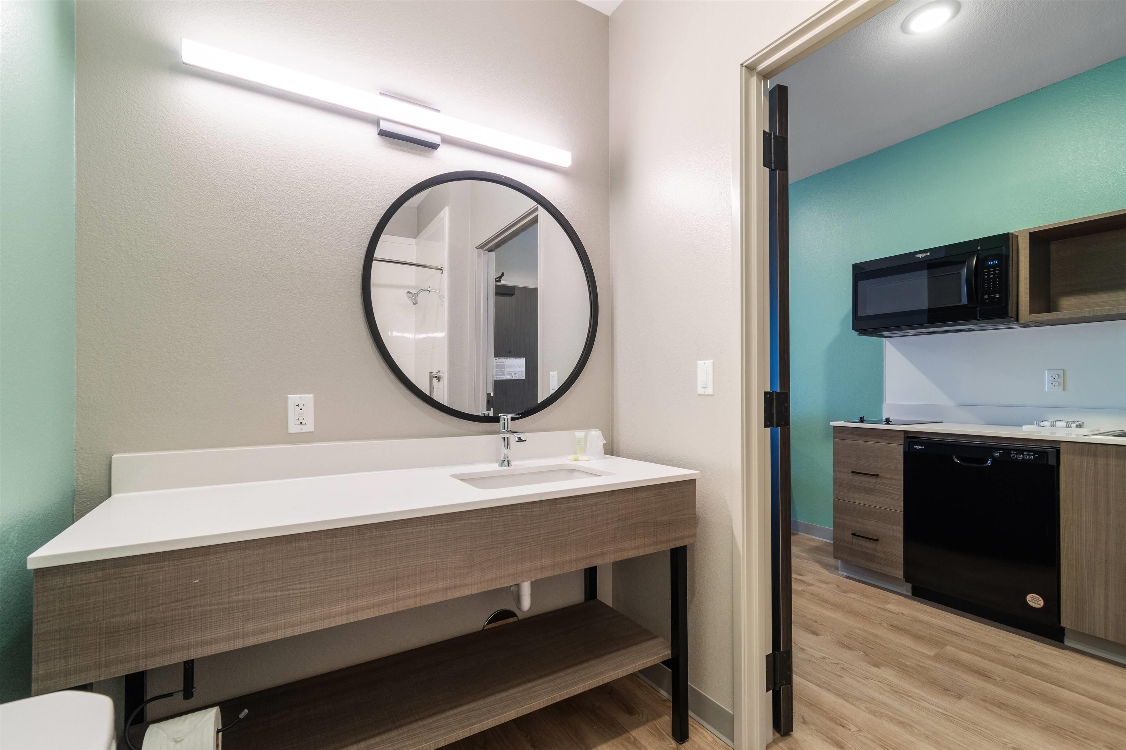 A modern bathroom with a white sink, a round mirror, and a black-framed mirror. There is a wooden cabinet below the sink and a black dishwasher to the right.