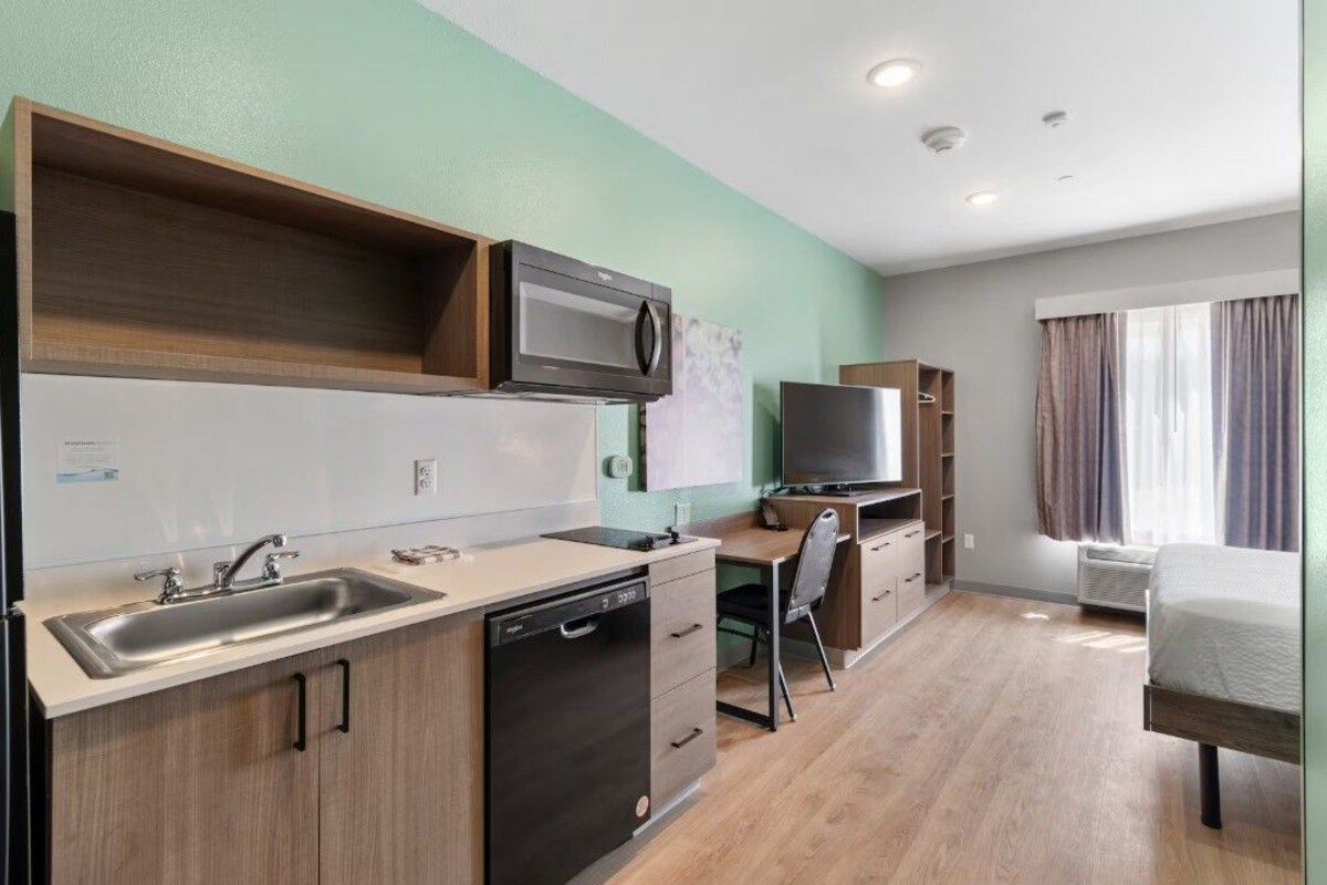 Una kitchenette compacta equipada con microondas, pileta y lavavajillas, un escritorio pequeño con silla, y una cama con cortinas.