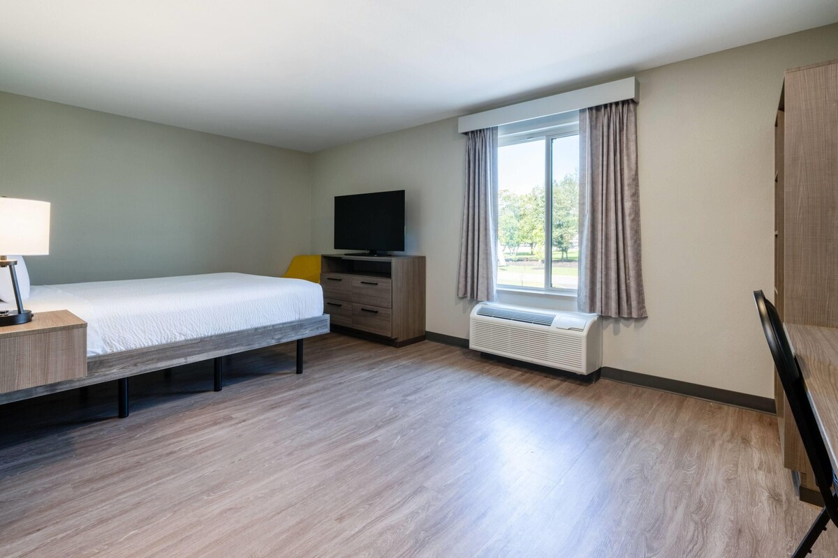 Une chambre d’hôtel avec un lit, une télévision, un bureau avec une lampe, une fenêtre avec des rideaux et un radiateur.
