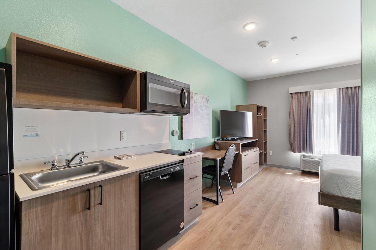 A compact kitchenette with a microwave, sink, and dishwasher, a small desk with a chair, and a bed in a room with light-colored walls and wooden flooring.