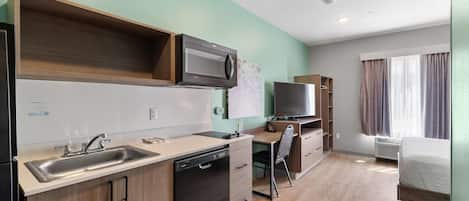 A compact kitchenette with a microwave, sink, and dishwasher, a small desk with a chair, and a bed in a room with light-colored walls and wooden flooring.