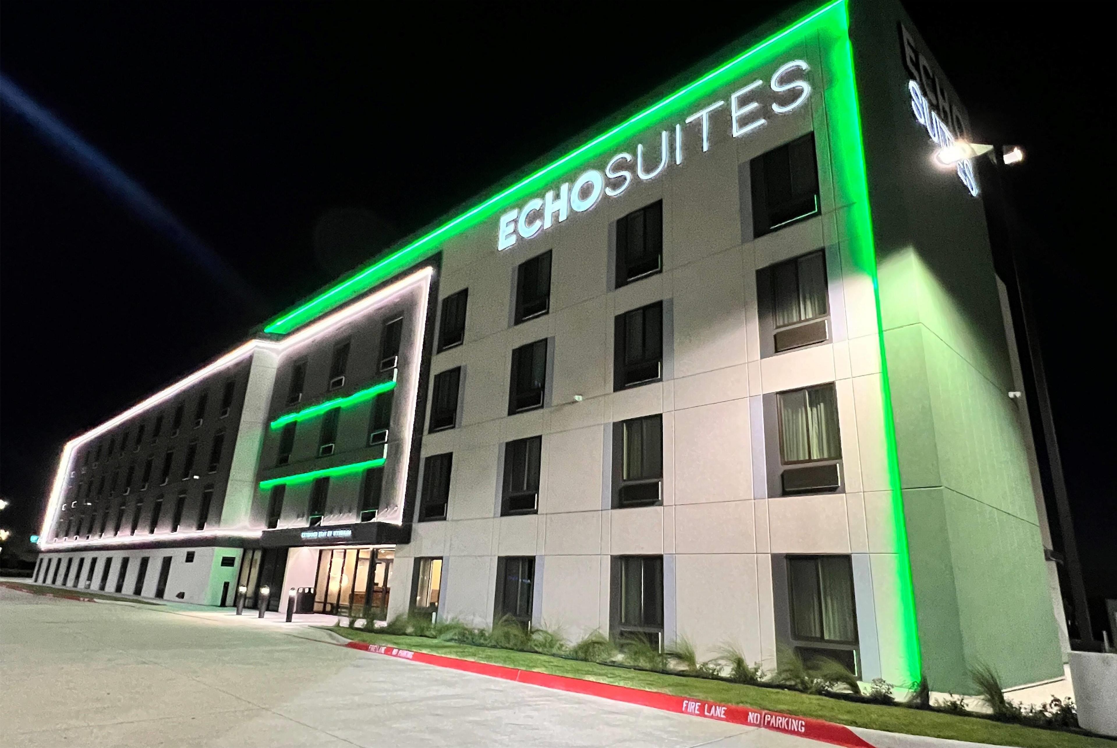A hotel named ECHOSUITES with a modern facade, illuminated green accents, and a fire lane in front.